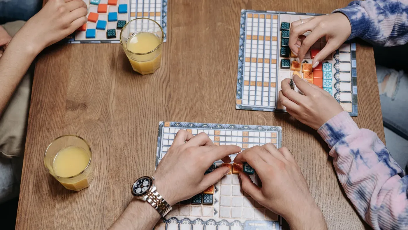 Hands around a wooden table placing colourful tiles on score sheets